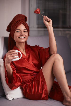 Model in a red bathrobe and a red towel on the sofa with a coffee mug and a lollipop. The concept of...