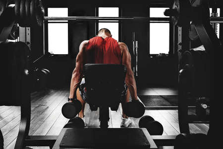 Young sports athlete fitness model in the gym shakes his biceps with dumbbells. Sports motivation, low key, high contrast. The concept of a healthy lifestyle, life movement, copy space.の写真素材