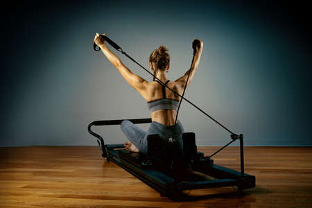 Young girl doing pilates exercises with a reformer bed. Beautiful slim fitness trainer on reformer gray background, low key, art light. Fitness conceptの写真素材
