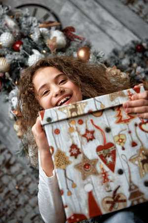 Funny smiling joyful child girl holds a Christmas present in his hand on a background of Christmas decorations and a Christmas tree. Waiting for a miracle, end Christmas and New Year. Portrait of a beautiful baby girlの写真素材