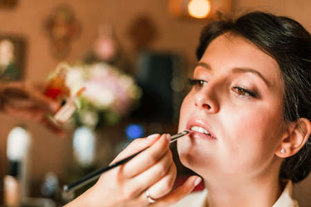 Makeup artist applies red lipstick . Beautiful woman face. Hand of make-up master, painting lips of young beauty model girl . Make up in processの写真素材