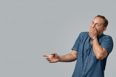 A man with scared face in a shirt shows with his hands on your text or your advertisement on a gray...