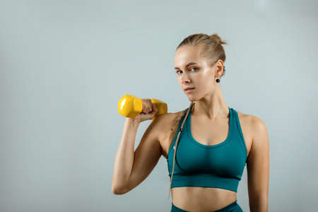 Beautiful fitness model doing exercises with dumbbells in hands on a gray background. Close-up. Woman posing on camera with dumbbells. Healthy lifestyle. Achieving goals, sports motivation. Copy spaceの写真素材