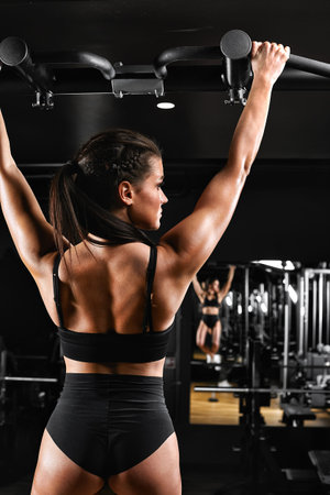 Sexy young fitness girl pulls up in the gym. Brunette fitness woman in black sportswear with perfect...