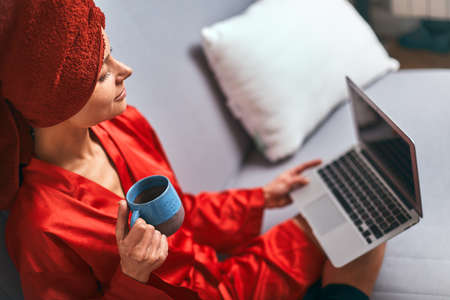 Young woman in red bathrobe and red towel on her head is standing in kitchen near table, drinking coffee and using laptop. Morning, girl after shower drinks tea and works on computer.Freelancer works at home.の写真素材