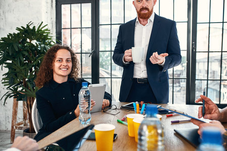 Couple of cheerful colleagues talking while standing at the office window with cups of coffee