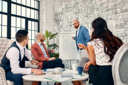 Confident businessman writing on flip charts presentation new project in boardroom at company meeting. Serious mentor auditor speaks with partners about business using board and graphs.の写真素材