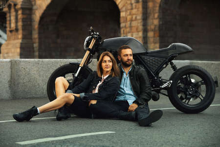 Beautiful biker couple in love are sitting on the road next to motorcycles in the cityの写真素材