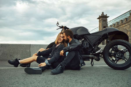 Beautiful biker couple in love are sitting on the road next to motorcycles in the cityの写真素材