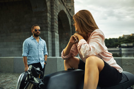 Romantic date on motorbike. Young woman sits on a motorcycle and looks at the man who comes to her. Couple in love with sunset under the bridge in the cityの写真素材