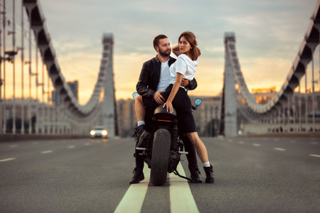 Young sexy woman hugging cute man in stylish black leather jacket, sitting on sports motorcycle on the bridge in the city on sunset and kissingの写真素材