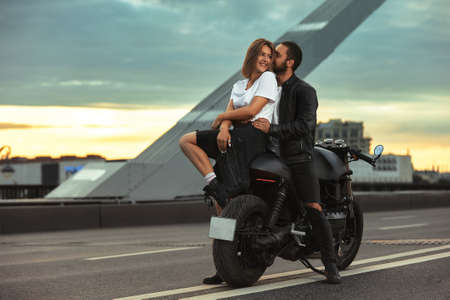 Young sexy woman hugging cute man in stylish black leather jacket, sitting on sports motorcycle on the bridge in the city on sunset and kissingの写真素材