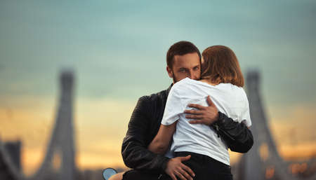 Young sexy woman hugging cute man in stylish black leather jacket, sitting on sports motorcycle on the bridge in the city on sunset and kissingの写真素材