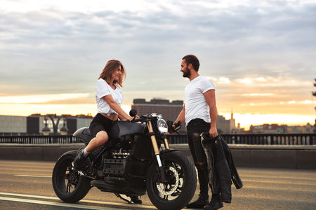 Romantic date on motorbike. Young woman sits on a motorcycle and looks at the man who comes to her. Couple in love with sunset on the bridge in the cityの写真素材