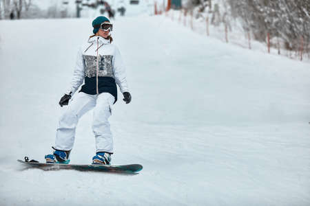 cheerful woman riding on snowboard in the mountains in winter. woman in white ski suitの写真素材