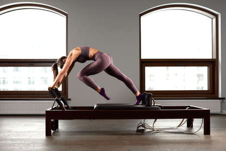 Young girl doing pilates exercises with a reformer bed. Beautiful slim fitness trainer on reformer gray background, low key, art light. Fitness conceptの写真素材