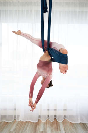 Cheerful sporty woman wearing top and leggings performing anti-gravity yoga exercise at spacious health club with panoramic windowsの写真素材