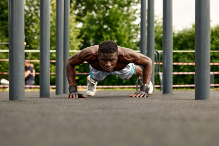 Muscular young man exercising at the sports field. African man looking to the side while doing horizontal bar exercises. Shirtless male model exercising outdoors.の写真素材