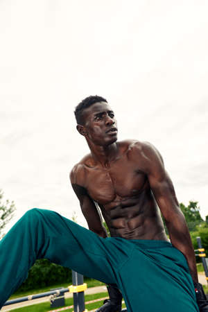 Muscular young man exercising at the sports field. African man looking to the side while doing horizontal bar exercises. Shirtless male model exercising outdoors.の写真素材