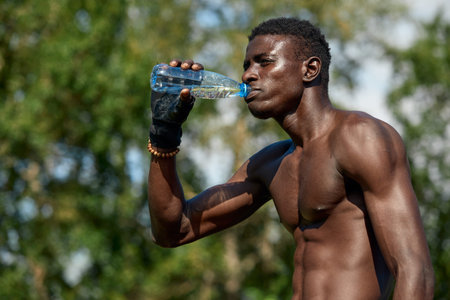 Young african american man drinks water while training in the park on the sports ground, outdoor...