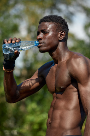 Young african american man drinks water while training in the park on the sports ground, outdoor workout concept, crossfitの写真素材