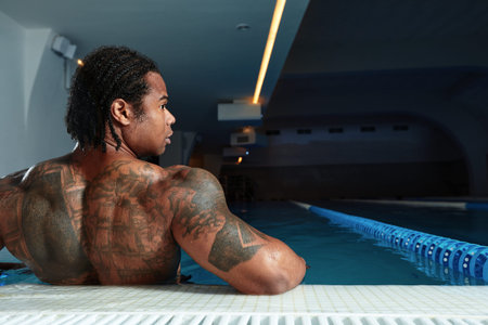 Young hot man resting at swimpool. Back view of guy sitting at water alon. Enjoy his time lonely in...