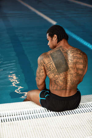 Young hot man resting at swimpool. Back view of guy sitting at water alon. Enjoy his time lonely in resort spa place. Strong powerful man.の写真素材
