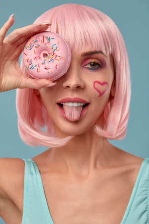 Portrait of a fashionable girl in a pink wig and with a pink donut in her hands keeps to the eye, shows tongueの写真素材