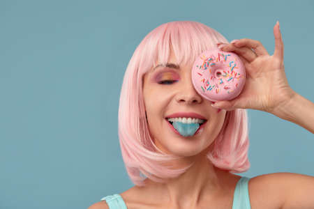Portrait of a fashionable girl in a pink wig and with a pink donut in her hands keeps to the eye, shows tongueの写真素材