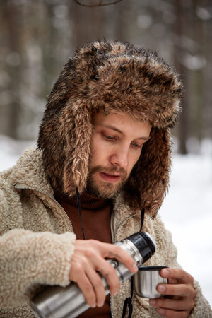 Handsome hiker man in warm clothes pours tea in mug from thermos in winter forest. Season concept. Selective focus.の写真素材
