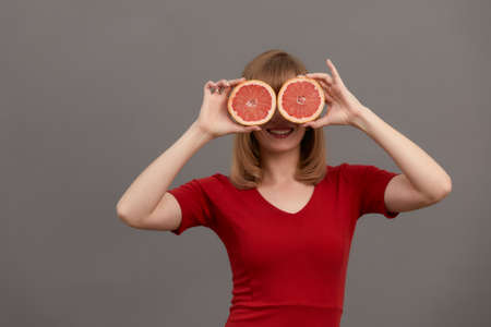 Beautiful woman with citrus fruits in hands at the eyes. Model with long hair hold cut grapefruit in hands as glasses isolated on gray. Skin care concept. Healthcare concept.の写真素材