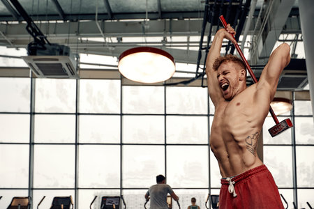 Strength concept. Male athlete using hammer during fitness workout. Fitness man beating rubber tire...