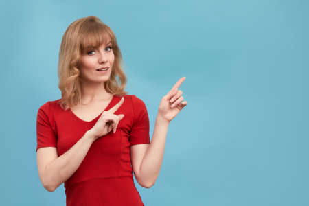 Happy surprised blonde young female smiling broadly at camera, pointing fingers away, showing something interesting and exciting on studio wall with copy space for your text or advertising contentの写真素材