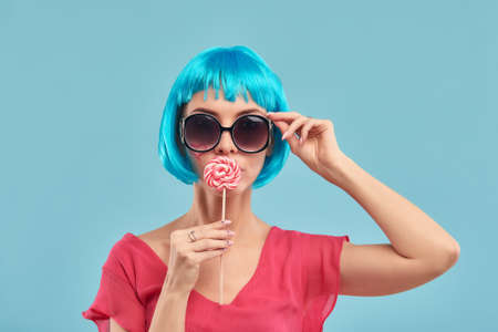 Young funky girl in blue wig and eyeglasses, holding lollipop candy, looking aside with dreamful smile as if making plans for party, isolated on blue backgroundの写真素材