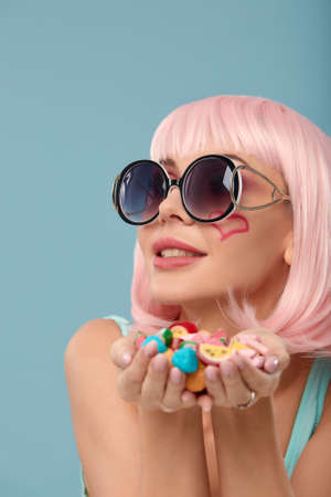Close up of a flirtatious female model on blue background wearing a pink wig holding different kinds of candies. Pretty glamorous woman pink hair charm sweets lifestyle enjoyment sugarの写真素材