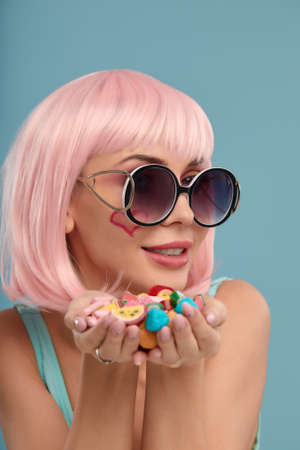 Close up of a flirtatious female model on blue background wearing a pink wig holding different kinds of candies. Pretty glamorous woman pink hair charm sweets lifestyle enjoyment sugarの写真素材