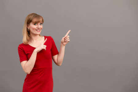 Happy surprised blonde young female smiling broadly at camera, pointing fingers away, showing something interesting and exciting on studio wall with copy space for your text or advertising contentの写真素材