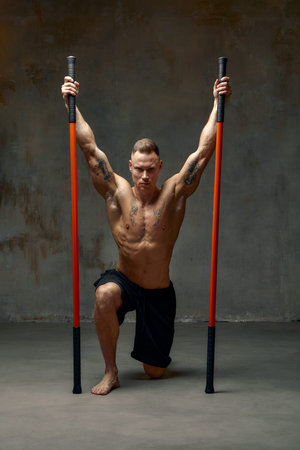 A fighter shirtless in black shorts, stands in a fighting artificially pose and holds a fight stick on studio background.の写真素材
