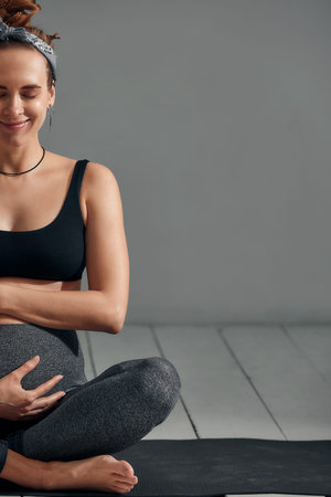 Young pregnant millennial woman sitting on the mat touches her belly after performing prenatal and...