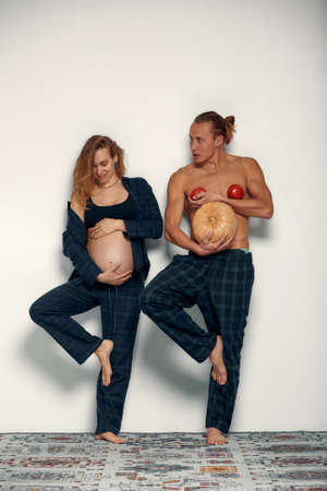 Funny pregnant girl with her husband play. Pregnant woman holds her belly and her husband holds a pumpkin instead of his belly and tomatoes at his chestの写真素材