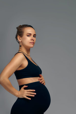 Cute pregnant belly isolated on gray. Side view of young pregnant woman embracing her abdomen with hands. Big belly on the third trimester of pregnancy close-up. Concept of pregnant life.の写真素材