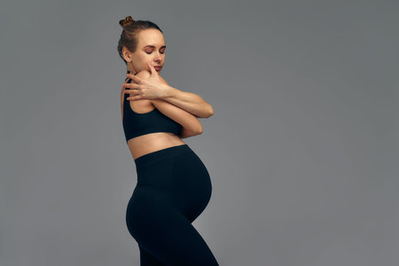 Cute pregnant belly isolated on gray. Side view of young pregnant woman embracing herself with...