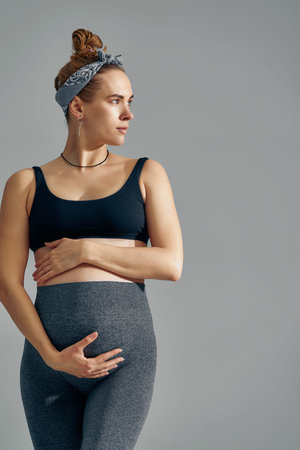 Cute pregnant belly isolated on gray. Side view of young pregnant woman embracing her abdomen with...