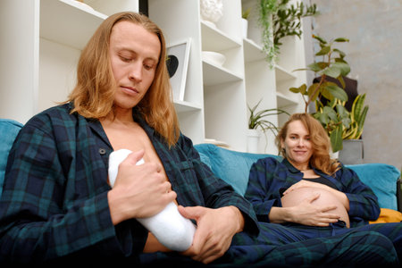 A husband massages his wifes feet and they sit together on the sofa in the living room. Pregnant...
