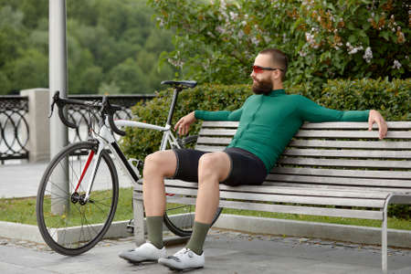 Stylish handsome guy cyclist rests on a wooden bench near bicycle in summer park.の写真素材
