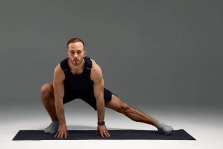 Attractive healthy young sports man stretching on fitness mat indoors, over gray wall backgroundの写真素材