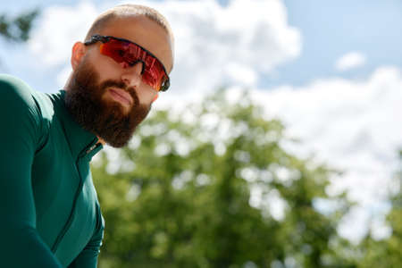 Close up shot of happy attractive young European man with beard wearing glasses and green sportswear looking at camera during evening ride in park on weekend.の写真素材