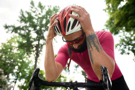 Tired male cyclist in sportswear, glasses and protective helmet walking with his bike in the forest to take a break after riding. Sky blue and forest in the background.の写真素材