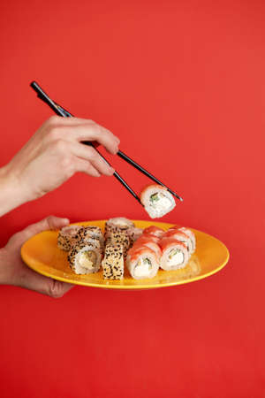 Close up cropped photo of female hand holds makizushi sushi roll served on plate traditional japanese food isolated on red background in studio. Copy space advertising mock up.の写真素材
