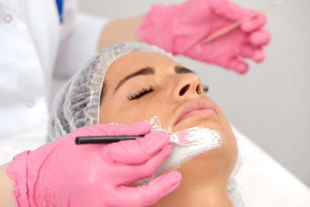 Beautician uses brush to apply white mask made of kaolin clay on half of womans face. Close up of young woman in white headband lying on soft towel in beauty salon. Concept of cosmetic care for faceの写真素材